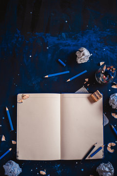 Open notepad with blank pages, crumpled paper balls, pencils, notepads and empty coffee cups on a dark background. Still life with writer workplace. Creative writing concept.