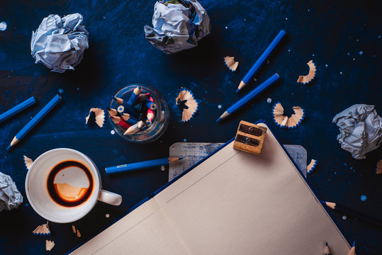 Open notepad with blank pages, crumpled paper balls, pencils, notepads and empty coffee cups on a dark background. Still life with writer workplace. Creative writing concept.