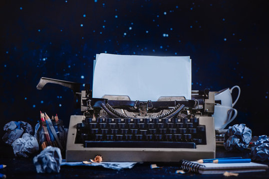Modern typewriter with sheets of paper, empty coffee cups, pencils and notepads on a dark background. Editing and copywriting workplace. Creative writing concept.