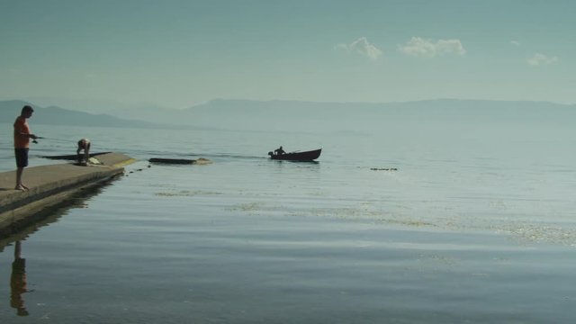 Man on the shore waves at passing by fisherman. Old traditional fishing boat along lake shore. Ohrid Lake Landscape on a summer sunny day.