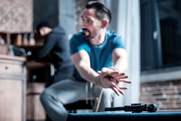 Fototapeta na wymiar Escaping. Frightened panic bearded man sitting with his hands tied and trying to take the gun while the criminal standing behind him