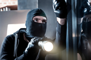 Breaking into. Dark-eyed professional masked burglar opening a window and holding a torch and...