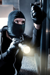 Thief. Dark-eyed professional masked burglar opening a window and holding a torch while breaking...