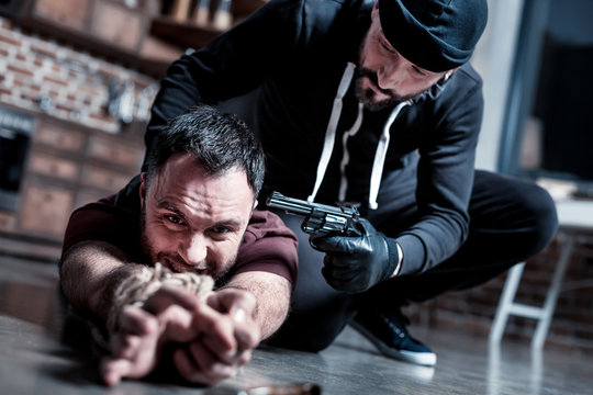 Trying to escape. Panic shocked bearded man lying on the floor with his hands tied and trying to escape while a killer sitting near him and holding a gun at his head