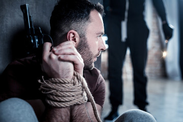 Hiding. Agitated panic bearded young man holding a gun and sitting with his hands tied while hiding...