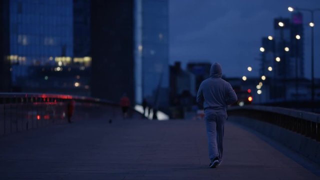 Wide shot of a hooded man walking along an empty bridge early in the morning, in slow motion
