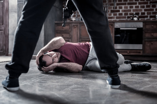 Victim. Frightened dark-eyed bearded man lying on the floor and covering his head with hands and a killer holding a pistol at his head