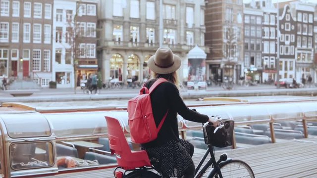 Female blogger rides bicycle along a river quay. Tourism. Caucasian girl in hat on a city bike. Side view slow motion.