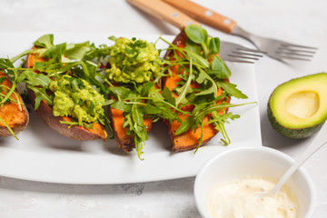 Baked halves of sweet potato with arugula and guacamole. White plate, light background, top view. Healthy vegan food concept.