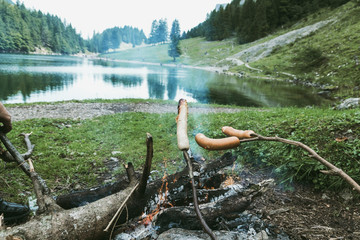 wooden sticks grilling barbecue sausages on fireplace in front of mountain lake