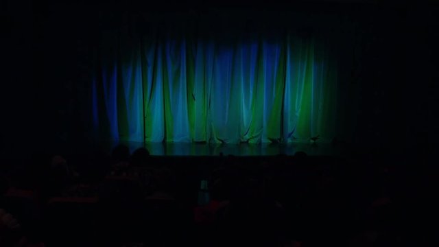 People, parents, the children in the auditorium before the show. Blue-green curtain. A theatrical production.