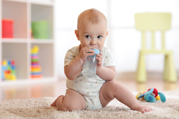 Pretty baby boy drinking water from bottle. Kid sitting on carpet in nursery at home. Smiling child...