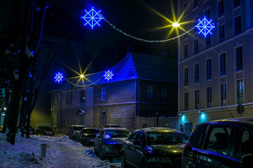Snowy christmas evening in city.  Illuminated blue buildings  and lot of cars in row.  Christmas decorations.