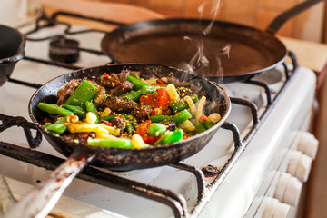 Red meat with vegetables cooking in an old dirty fry pan, steam rising in sunlight