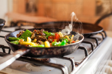 Red meat with vegetables cooking in an old dirty fry pan, steam rising in sunlight