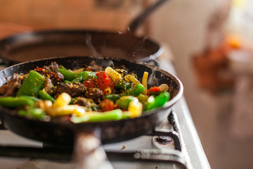 Red meat with vegetables cooking in an old dirty fry pan, steam rising in sunlight