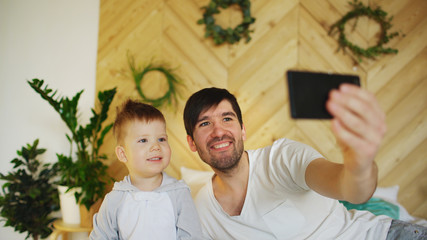 Fototapeta premium Young smiling father with son on bed taking selfie photo with smartphone camera