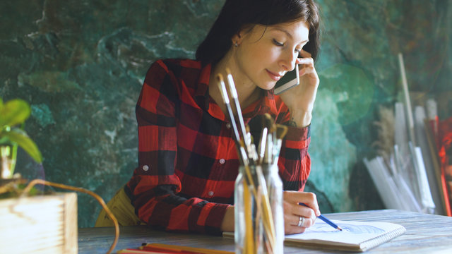 Young woman artist painting scetch on paper notebook with pencil and talking phone indoors