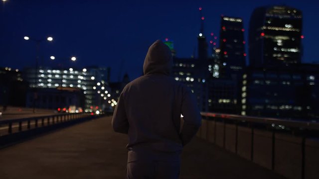 Wide angle shot of camera following a hooded male jogging towards city lights, in slow motion
