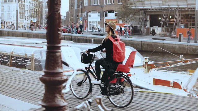 Cute European girl rides bicycle along river quay. Touristwoman in hat on a city bike in Amsterdam. Real time side view.