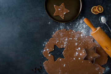 Christmas gingerbread star cookie cutter on gingerbread dough with cookie cutout, star shaped cookie on plate, teaspoon of flour, dried orange slices and rolling pin, dark background, top view.