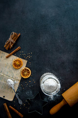 Food preparation. Old recipe, teaspoon, cookie cutter, rolling pin, mason jar with flour and ingredients (cinnamon sticks, dried orange slices) on dark modern table from above. Top view.
