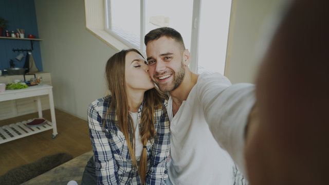 POV of Young smiling and beautiful couple take selfie portrait on smartphone camera and kiss while sitting on bed in the morning