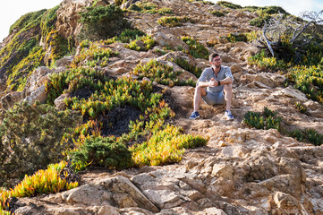 Young and handsome man sitting on a rock after a walk among poor vegetation and stones
