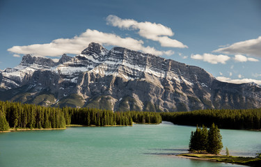 Two Jack Lake with Mount Rundle. A sunny day with a few clouds over the mountain. The lake is a turquoise color and trees are a rich green. There is snow on mount Rundle.