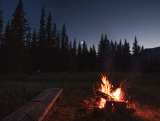 Campfire under the out of focus moon. There is a wooden bench beside the campfire with a hot dog / marshmallow roasting stick laying on the bench. There is a silhouette of the trees in the background.