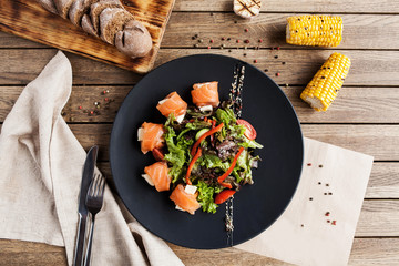 salad with vegetables, cheese and fresh salmon rolls on a black plate with a bread board and corn on a wooden table - 187334635