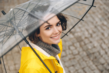 Amazing portrait of young woman in yellow coat standing under transparent umbrella with broad sincere smile