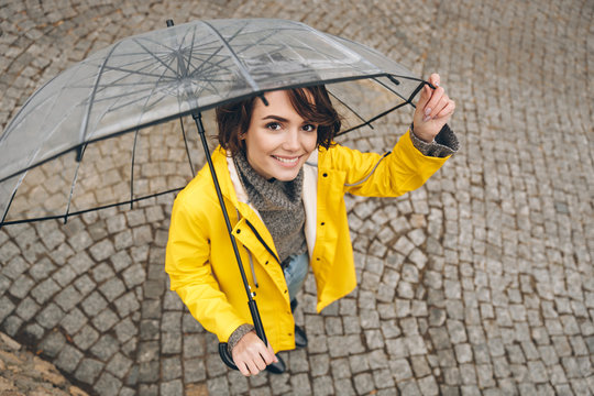 Shot From Above Of Wonderful Woman In Yellow Raincoat Being Happy While Walking Under Big Transparent Umbrella