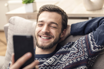 Obraz premium Cheerful young man holding mobile phone and smiling at home.
