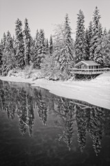 Black and white winter snow scene of cabin and snow covered trees reflecting in frozen river