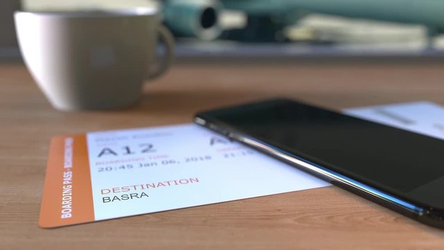 Boarding pass to Basra and smartphone on the table in airport. Travelling to Iraq conceptual animation
