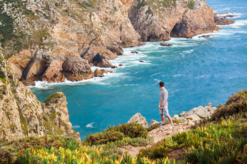 Stylish, young man standing at the very edge of the rock and staring into the distance (the place where the earth ends and the sea begins, Cabo da Roca (Cape Roca), Sintra, Portugal)