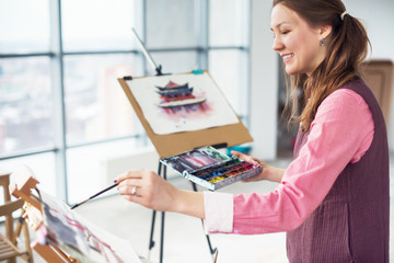 Portrait of a young woman painter drawing with watercolor palette on paper using easel.