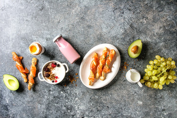 Breakfast set with smoked salmon on crispy breadsticks, yogurt, smoothie, avocado, muesli, milk, berries and green grape over blue texture background. Top view, copy space. Food background