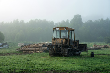 Fototapeta premium The caterpillar tractor stands in the morning fog on the background of logs and trees. Green grass.