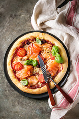 Traditional pizza with smoked salmon, cheese, tomatoes and basil served on black plate with kitchen towel over old dark metal background. Top view with space. Rustic style