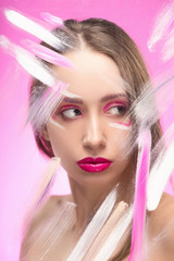 The beautiful girl with a vanguard make-up behind the glass covered with paint dabs on a pink...