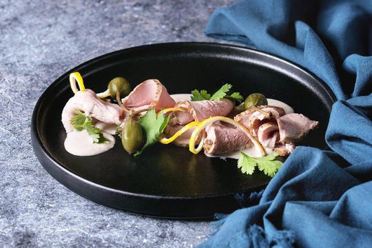 Vitello tonnato italian dish. Thin sliced veal with tuna sauce, capers and coriander served on black plate with blue textile over blue texture background. Close up