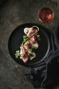 Vitello tonnato italian dish. Thin sliced veal with tuna sauce, capers and coriander served on black plate with glass of rose wine over old dark metal background. Top view, space