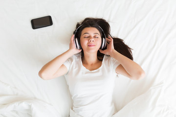Young woman lying on sofa and listening to music, relaxing with eyes closed on white bed at home at the morning