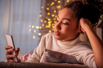 Portrait of pretty calm african girl using cellphone while relaxing at home. Light garland is on background