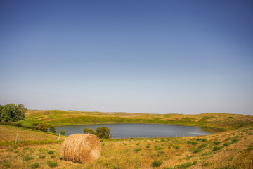 One round straw bale by a large pond of water surrounded by a pasture fence in a rural agricultural summer countryside landscape