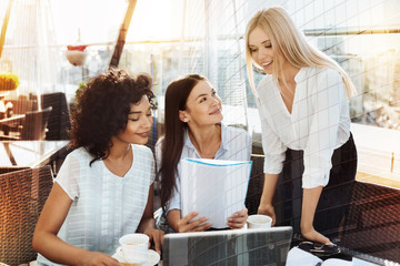 Have a look. Pretty young woman keeping smile on her face and bowing head while leaning arms on table