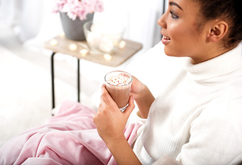 Charming african girl is enjoying hot cocoa with marshmallows at home. She is relaxing and smiling. Focus on cup in her hands. Copy space