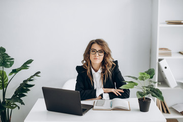 Serious business woman in a glasses and in suit, with a laptop sits at her workplace in a bright...